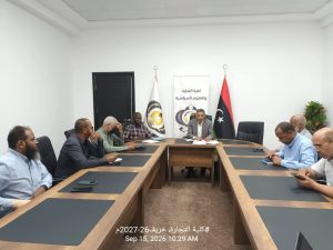 8435F9BA-38C3-47F7-B7B3-B80CE323B806 - جامعة سبها | Sebha University Business meeting in a bright conference room with several men seated around a long wooden table; flags and a banner with Arabic text behind the central figure.