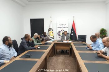 Business meeting in a bright conference room with several men seated around a long wooden table; flags and a banner with Arabic text behind the central figure.