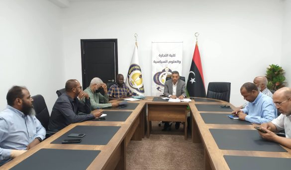Business meeting in a bright conference room with several men seated around a long wooden table; flags and a banner with Arabic text behind the central figure.