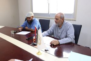 Two men sit at a conference table reviewing documents; flag flags and papers in front, window behind.