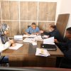 Four people sit around a wooden conference table in an office, reviewing documents; woman in a patterned hijab writes notes while three men discuss papers (one wearing a mask).
