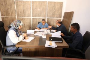 8ED75857-FC84-4B28-9967-63C09344CD16 - جامعة سبها | Sebha University Four people sit around a wooden conference table in an office, reviewing documents; woman in a patterned hijab writes notes while three men discuss papers (one wearing a mask).