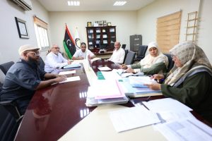 Six people sit around a long conference table reviewing documents; Libyan and another flag stand behind them in a formal meeting room.