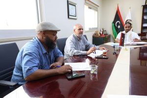 Three men sit at a long conference table reviewing documents; flags and a certificate are on the wall behind them.