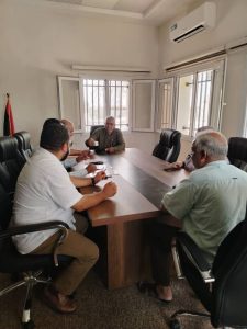 Six men sit around a rectangular conference table in a meeting room, one man in the center holding a cup and speaking to others.