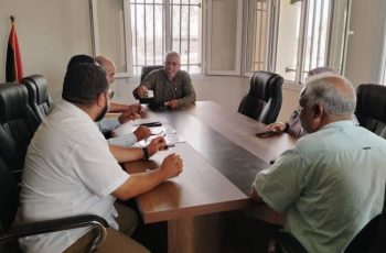 Six men sit around a rectangular conference table in a meeting room, one man in the center holding a cup and speaking to others.