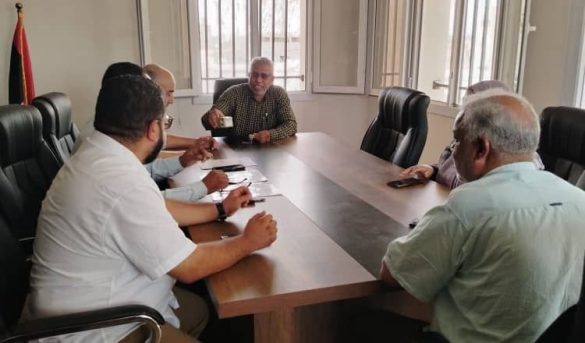 Six men sit around a rectangular conference table in a meeting room, one man in the center holding a cup and speaking to others.