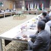 Formal conference room with attendees seated around a long U-shaped table, flag banners behind and a central floral arrangement in view.