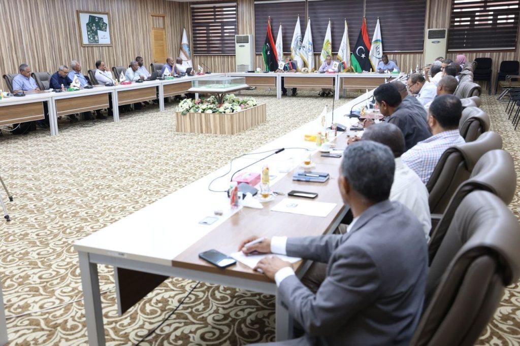 94577889-4CB9-4DAD-A128-7E972116E4CB - جامعة سبها | Sebha University Formal conference room with attendees seated around a long U-shaped table, flag banners behind and a central floral arrangement in view.