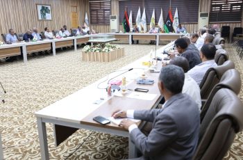 Formal conference room with attendees seated around a long U-shaped table, flag banners behind and a central floral arrangement in view.