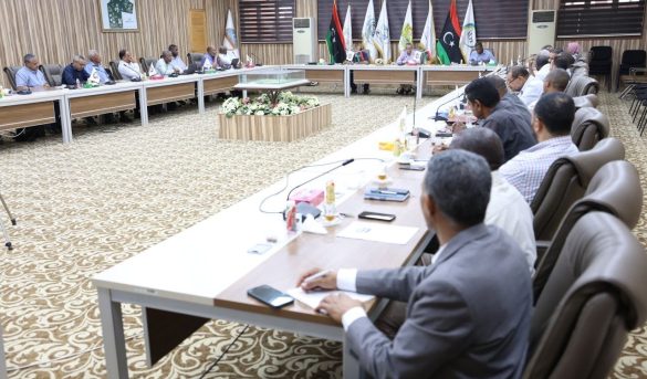 Formal conference room with attendees seated around a long U-shaped table, flag banners behind and a central floral arrangement in view.