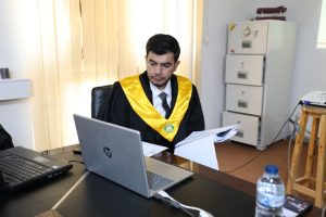 Man in a black academic gown with a yellow ceremonial sash sits at a desk, reviewing documents with a laptop open in front of him.