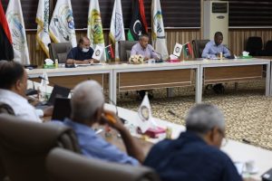 Formal meeting in a conference room with officials seated along a long table, Libyan flags and organizational banners visible in the background.