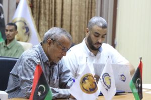 9EE1C8FD-112B-4060-9110-328ACE82FECD - جامعة سبها | Sebha University Two men sign documents at a formal meeting, with Libyan flags on the table in front of them