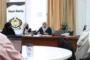 9EEEA77A-AFC1-41ED-9A47-FEE735932DE5 - جامعة سبها | Sebha University Four women in hijabs sit around a long conference table in a meeting room, taking notes and using laptops, with a banner featuring Arabic text in the background.