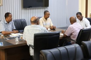 Five men in a boardroom seated around a wooden conference table, taking notes and chatting.]​','Meeting in progress: five professionals around a table with notebooks, pens, and tea cups.