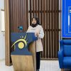 Woman in hijab speaking at a podium with a blue and gold sun-logo, microphone nearby in a formal setting