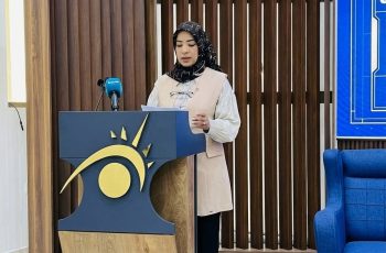 Woman in hijab speaking at a podium with a blue and gold sun-logo, microphone nearby in a formal setting