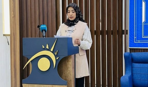 9F427FDB-FDD4-4576-A096-8C04D913F3A4 - جامعة سبها | Sebha University Woman in hijab speaking at a podium with a blue and gold sun-logo, microphone nearby in a formal setting