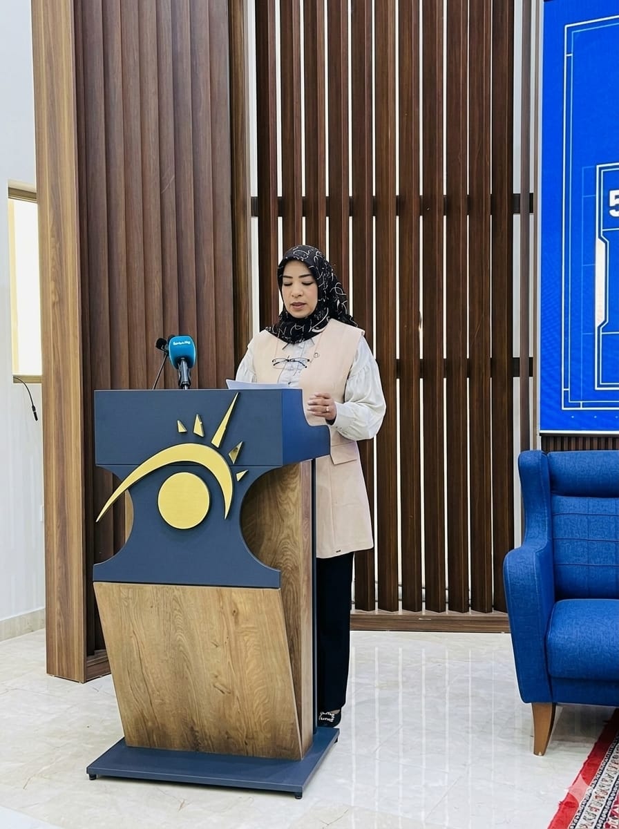 Woman in hijab speaking at a podium with a blue and gold sun-logo, microphone nearby in a formal setting