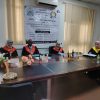 Four academics in red and black academic gowns sit at a long conference table in a meeting room with a large banner in Arabic on the wall behind them.