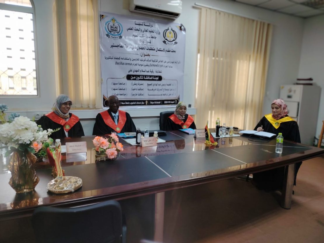 Four academics in red and black academic gowns sit at a long conference table in a meeting room with a large banner in Arabic on the wall behind them.
