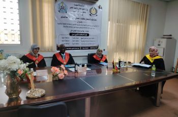 Four academics in red and black academic gowns sit at a long conference table in a meeting room with a large banner in Arabic on the wall behind them.