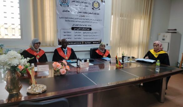 A01B801D-8DCA-4AF9-811D-A72D6F993ABD - جامعة سبها | Sebha University Four academics in red and black academic gowns sit at a long conference table in a meeting room with a large banner in Arabic on the wall behind them.