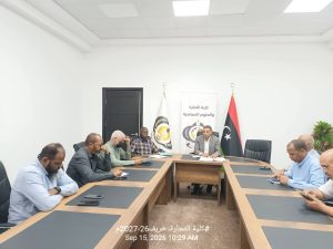A0EF8D7E-E95D-43A9-8F46-3D7AD834CB4B - جامعة سبها | Sebha University People sit around a large conference table in a bright room; banners and a Libyan flag visible in the background, suggesting a formal meeting or signing event.