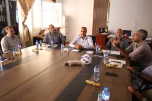 A1FB14A6-D702-4C60-80B5-107F67D206BD - جامعة سبها | Sebha University Six men sit around a large conference table in a bright office, engaged in a discussion with water bottles and papers on the table nearby, windows open in the background.