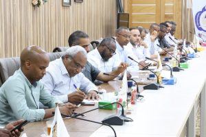 Group of men seated along a long conference table, each with microphones, documents, and beverages, in a formal meeting room