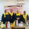 Four men in black academic robes with yellow hoods stand in front of a banner at a formal event, flags and snacks on the table in front of them.