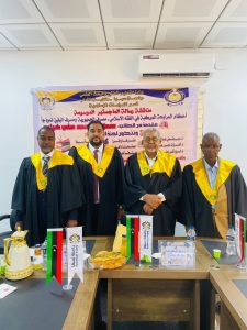 A5E48D49-9DF4-4B48-8E4C-F0B433855192 - جامعة سبها | Sebha University Four men in black academic robes with yellow hoods stand in front of a banner at a formal event, flags and snacks on the table in front of them.
