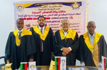 Four men in black academic robes with yellow hoods stand in front of a banner at a formal event, flags and snacks on the table in front of them.