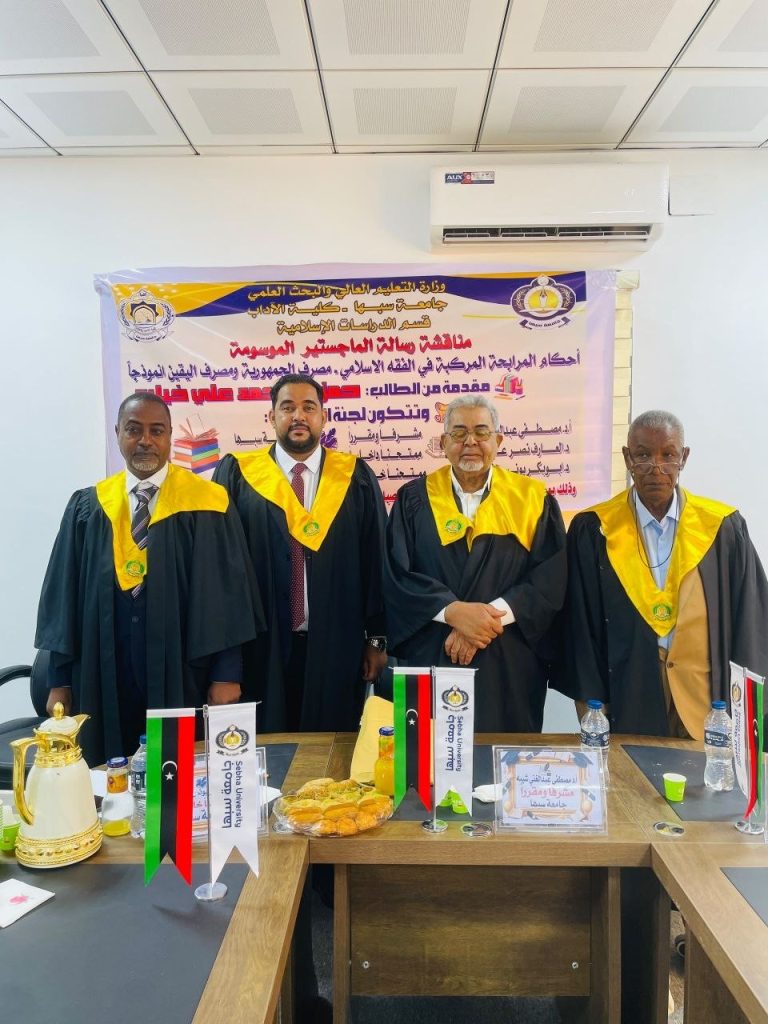 Four men in black academic robes with yellow hoods stand in front of a banner at a formal event, flags and snacks on the table in front of them.