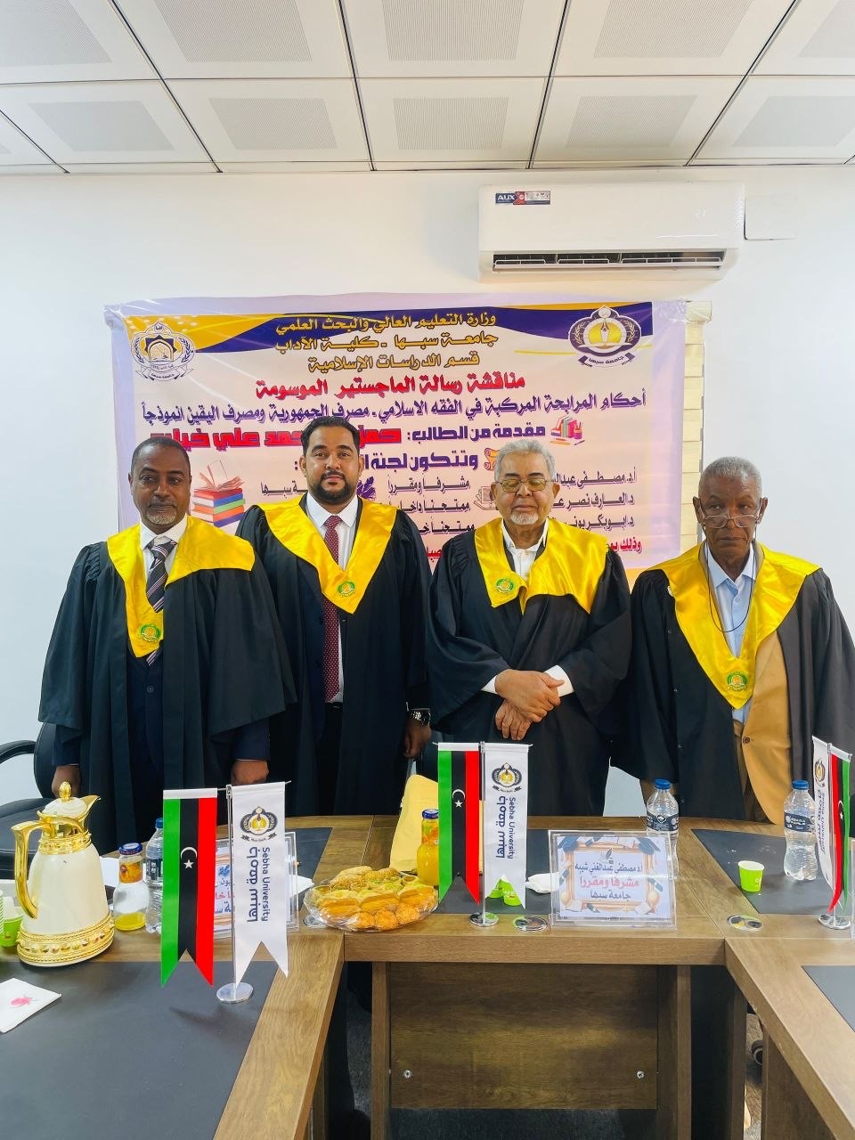 Four men in black academic robes with yellow hoods stand in front of a banner at a formal event, flags and snacks on the table in front of them.