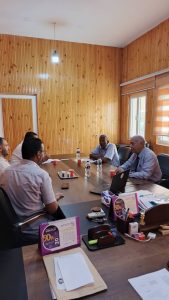 A business meeting in a wood-paneled room with five men seated around a long table, laptops, papers, and water bottles visible on the table.