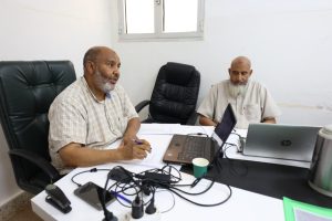 Two older men sit at a cluttered office desk with laptops and papers, engaged in a discussion.