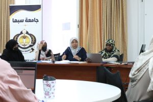 A9CF5C9C-8FEF-4949-8473-1DF05016A865 - جامعة سبها | Sebha University Group of women in hijabs seated around a conference table during a university meeting; banner with Arabic text and crest on the left.