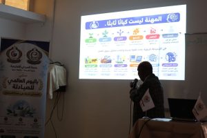 AAA1FEBD-B5DB-4004-842A-2A734F7B6C44 - جامعة سبها | Sebha University Man with a microphone presenting a projector slide in Arabic, featuring a colorful infographic and a standing banner beside him.