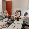 Three colleagues in a meeting around a cluttered table with papers, water bottles, and coffee pots.