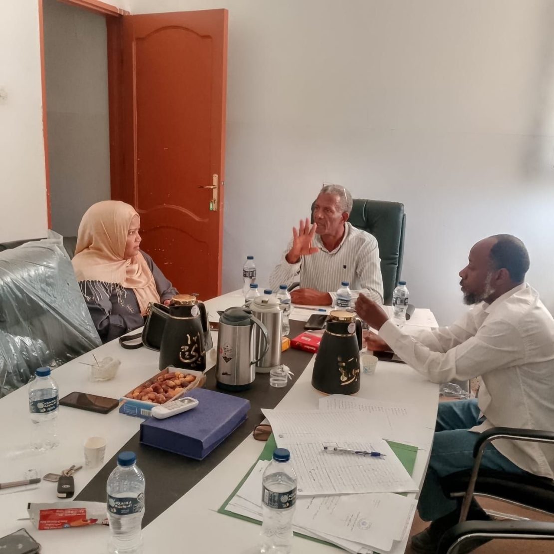 Three colleagues in a meeting around a cluttered table with papers, water bottles, and coffee pots.