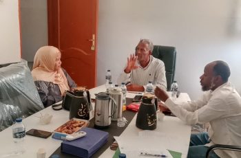 Three colleagues in a meeting around a cluttered table with papers, water bottles, and coffee pots.