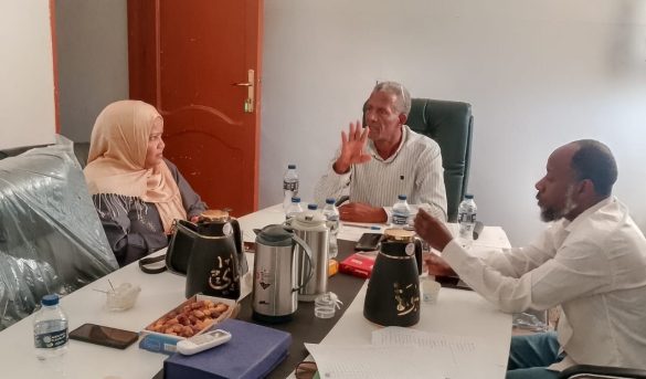 Three colleagues in a meeting around a cluttered table with papers, water bottles, and coffee pots.