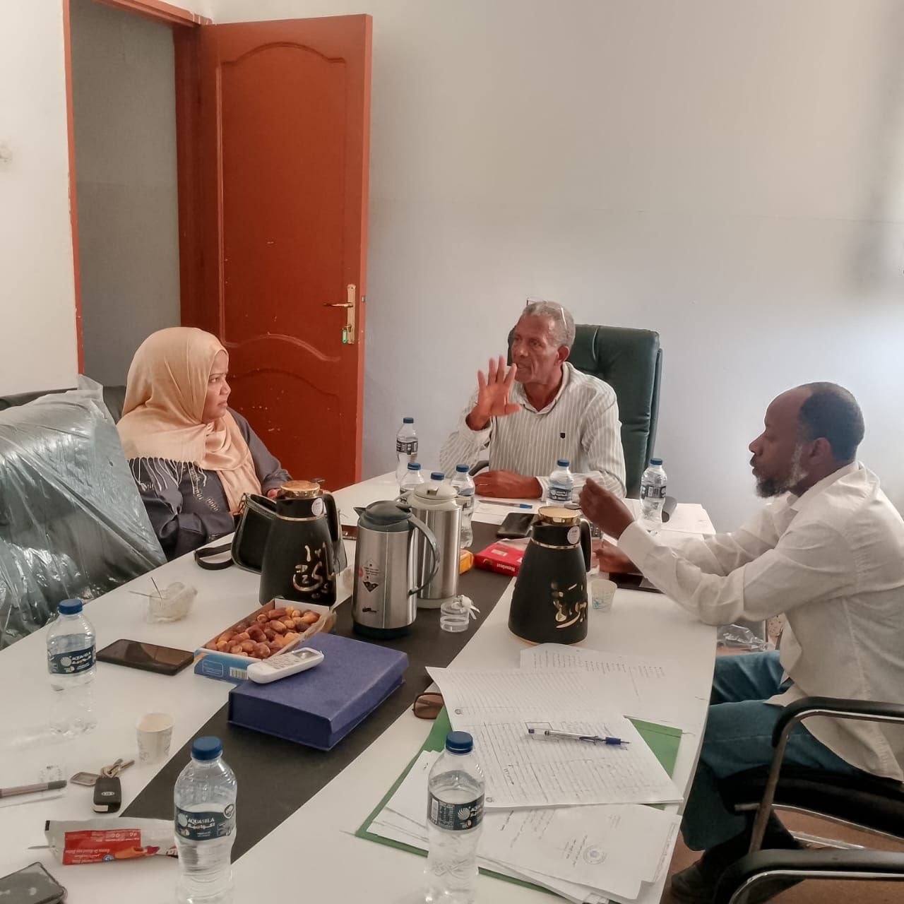 Three colleagues in a meeting around a cluttered table with papers, water bottles, and coffee pots.