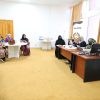 Group of women in hijabs seated around tables in a meeting room, with a banner and computer at the front.