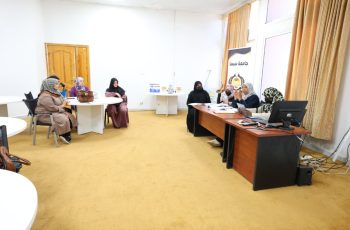 Group of women in hijabs seated around tables in a meeting room, with a banner and computer at the front.