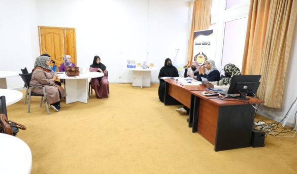 BA445FE9-E0A8-4AE0-9CDB-920D4D3542BB - جامعة سبها | Sebha University Group of women in hijabs seated around tables in a meeting room, with a banner and computer at the front.