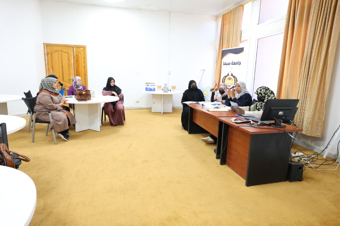 Group of women in hijabs seated around tables in a meeting room, with a banner and computer at the front.