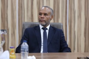 BECE8518-53FF-47AA-A687-21C5279E5627 - جامعة سبها | Sebha University A middle-aged man in a navy suit and tie sits at a conference table in a formal office setting, looking off to the side.